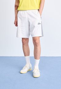 Bijele atletske kratke hlače s elastičnim pojasom, bočnim džepovima i crnim Adidas logotipom. U kombinaciji s laganim tenisicama i bijelim čarapama do gležnja.
