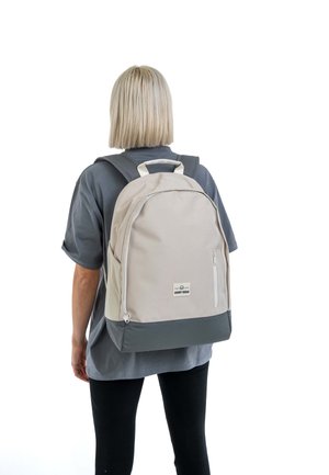 Beige fabric backpack with gray bottom, rectangular shape, front zip pocket, and dual shoulder straps, featuring a top handle and logo patch.