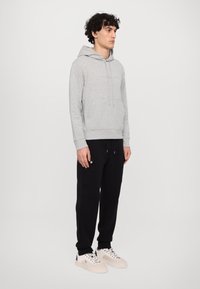 Felpa grigia chiaro con tasca frontale, pantaloni jogger neri con tasca laterale e sneakers crema con dettagli neri, mostrati su un modello rivolto a sinistra.