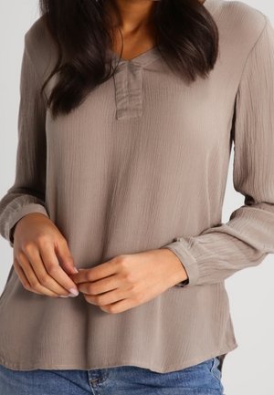 Femme portant un chemisier taupe texturé à manches longues avec une patte de boutonnage en V, associé à un jean en denim bleu, mains délicatement croisées.