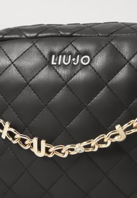 Bolso acolchado negro con una correa de cadena dorada y el nombre de la marca "LIU-JO" grabado en la parte frontal. Superficie texturizada con un acabado brillante.
