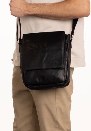Borsa a tracolla in pelle nera con chiusura a pattina, tracolla regolabile e texture liscia, tenuta da una persona su uno sfondo neutro.