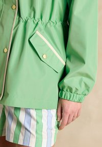 Zaļš ūdensnecaurlaidīgs mētelis ar elastīgiem piedurkņu un jostas galiem, ar krūšu kabatu, kurai ir zelta poga un ziloņkaula apdare. Zem tā ir svītrainas šorti.