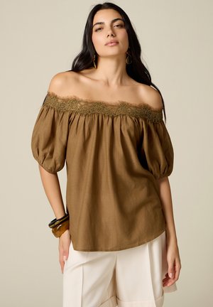 Donna con lunghi capelli scuri che indossa una blusa marrone a spalle scoperte con bordo in pizzo e pantaloni bianchi, accessoriata con bracciali e orecchini.