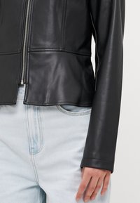 Black leather fitted jacket with a high collar, silver zipper, and smooth texture. Paired with light blue denim jeans.