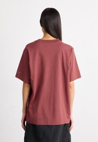 Kvinna med långt mörkt hår som bär en lös kortärmad mörkröd t-shirt och svart kjol, stående mot en enfärgad vit bakgrund.