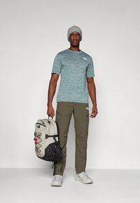 Zelena kratka majica z meliranim vzorcem, olive hlače s cargo žepi, siva kapa in bež/črni nahrbtnik z več žepi in nastavljivimi trakovi.