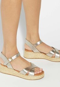Feet wearing silver metallic wedge sandals with crisscross straps and buckle, featuring woven textured platform soles.