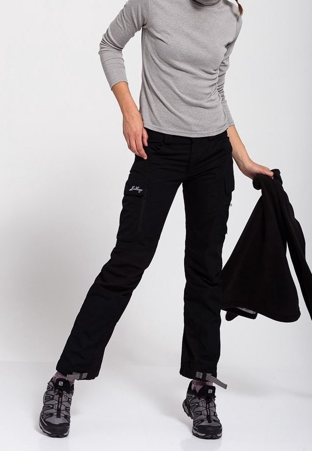 Personne portant une chemise grise à manches longues, un pantalon cargo noir et des chaussures de randonnée, tenant une veste noire dans une main.