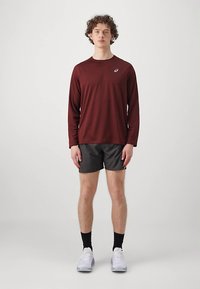 Junger Mann steht mit den Armen neben dem Körper, trägt ein burgunderfarbenes Langarmshirt, schwarze Shorts, schwarze Socken und weiße Turnschuhe vor einem neutralen Hintergrund.