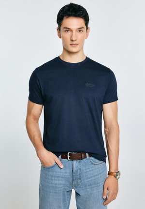 Granatowy t-shirt z krótkim rękawem wykonany z miękkiej, gładkiej tkaniny; posiada subtelne haftowane logo na piersi. Noszony z jasnymi, dżinsowymi spodniami.