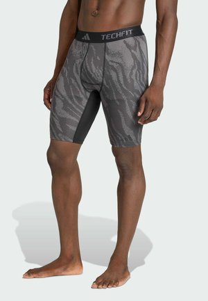 Buste et jambes masculins portant un short de compression Adidas Techfit gris à motifs, debout pieds nus sur un fond clair.