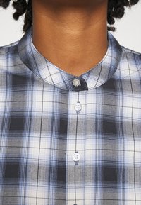 Camicia a quadri con colletto rotondo, caratterizzata da motivi blu, neri e bianchi. Chiusura con due bottoni, tra cui un bottone decorativo in perla.