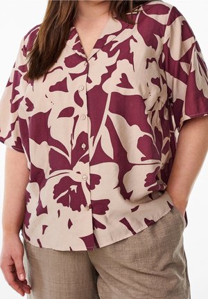 Kvinde iført beige og mørkerød blomstret kortærmet skjorte med knapper og lysebrune bukser, hånden i lommen, mod hvid baggrund.