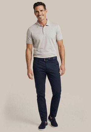 Lachende man staand, gekleed in een lichtgrijs poloshirt, donkerblauwe broek, donkerblauwe riem en donkerblauwe instapschoenen tegen een lichtgrijze achtergrond.