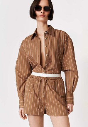 Kobieta ubrana w brązową koszulę w cienkie paski z guzikami, do niej dopasowane krótkie spodenki oraz biały pasek, uzupełniona dużymi ciemnymi okularami przeciwsłonecznymi.