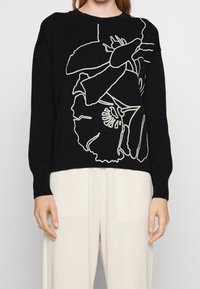 Kobieta w czarnej bluzie z abstrakcyjnym białym wzorem kwiatowym i kremowych, szerokich spodniach, stojąca na jednolitym tle.