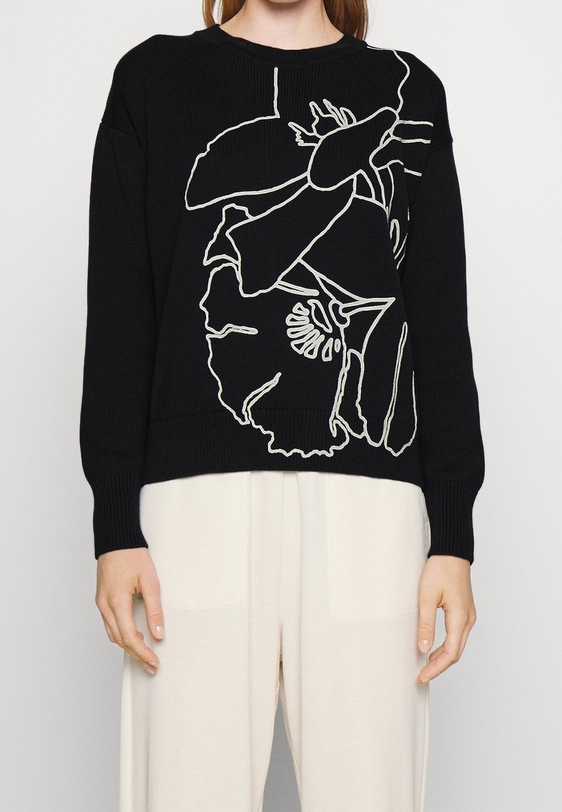 Kobieta w czarnej bluzie z abstrakcyjnym białym wzorem kwiatowym i kremowych, szerokich spodniach, stojąca na jednolitym tle.