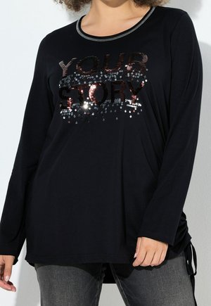 Zwarte top met lange mouwen en pailletten tekst "YOUR STORY", gedragen door een persoon met krullend haar, gecombineerd met donkergrijze jeans.