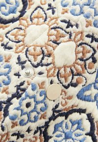 Tekstil med blomstermønster i blå, brun og cremede nuancer. Har en stofstruktur med sølvknapbeslag og indviklede design detaljer.