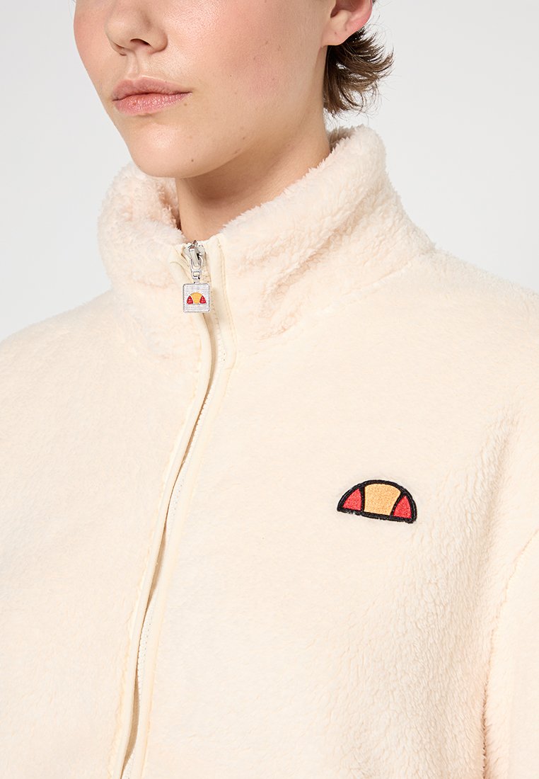 Veste zippée en polaire de couleur crème clair, dotée d'un col montant et d'un logo multicolore brodé sur la poitrine. Surface douce et texturée.