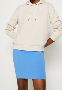 Sudadera beige con cordones y logo en relieve, combinada con una falda azul ajustada. Tejido suave con un diseño casual y detalles mínimos.