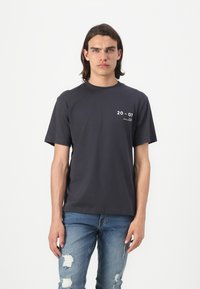 Mørk grå bomull t-skjorte for menn med korte ermer, rund hals og hvit trykk "20 - 07" på venstre brystområde. Brukt med falmede blå ripped jeans.