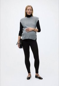 Έγκυος γυναίκα που φοράει μαύρο ζιβάγκο και κολάν με γκρι αμάνικο πουλόβερ, κρατώντας μαύρη τσάντα και αγγίζοντας την κοιλιά της.