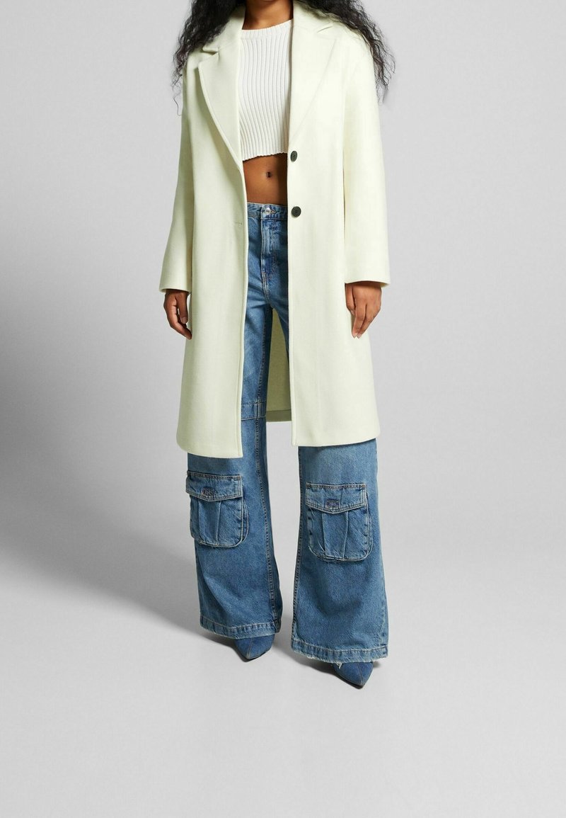 Lång krämfärgad kappa med slagkrage, två knappar, i kombination med högmidjade, vida blåjeans med cargofickor. Släta texturer.