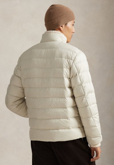 Šviesiai smėlio spalvos pūkinė striukė, su siuvinėtu raštu ir aukštu apykakliu. Yra ribuoti rankogaliai ir minkšta tekstūra. Dėvima su rudu megztu kepure.