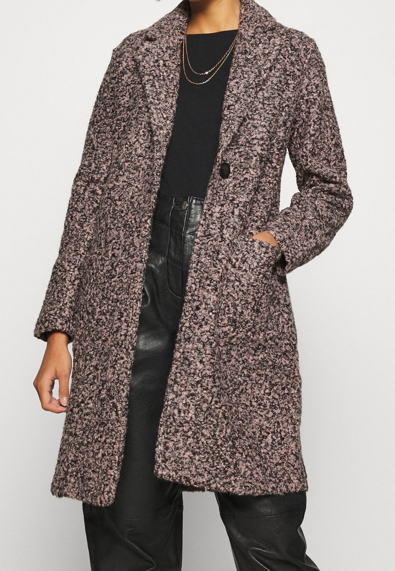 Mujer con abrigo de lana texturizado en marrón y negro sobre camisa negra y pantalones de cuero negros de talle alto, con una mano en el bolsillo del abrigo.