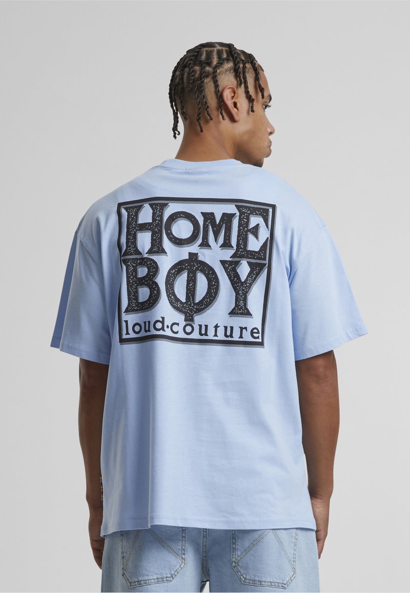 Man with braided hair wearing a light blue T-shirt that reads "HOME BOY loud.couture" in large black letters on the back.