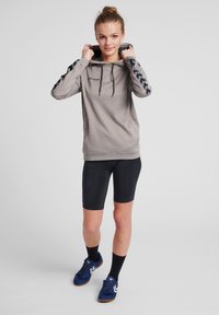 Felpa grigia con accenti a chevron neri, cappuccio con cordoncino, tessuto testurizzato. Abbinata a pantaloni corti neri e scarpe da atletica blu.