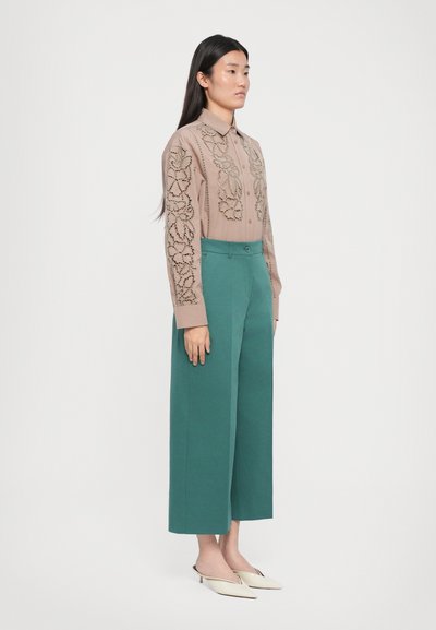 Beige Hemdbluse mit floralem刺绣 an den Ärmeln, kombiniert mit grünen, weiten Hosen und weißen Mules, vor einem weißen Hintergrund stehend.