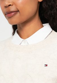 Crème kabelgebreide trui met een wit overhemd eronder. Bevat een klein meerkleurig logo op het linkerborstgebied.