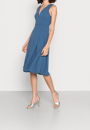 Woman in sleeveless blue midi dress with pleats and silver high-heeled sandals standing against a plain light background.