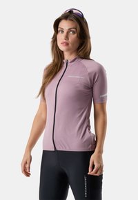 Kobieta ubrana w bladoróżową, krótkorękawową koszulkę kolarską i czarne spodenki kolarskie z logo Swedemount, stojąca na jednolitym tle.