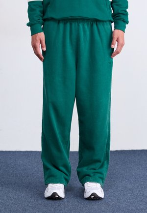 Pantalons de survêtement en mélange de coton vert avec une taille élastique et des poches latérales, présentant une coupe décontractée et des détails cousus. Associés à des baskets blanches.