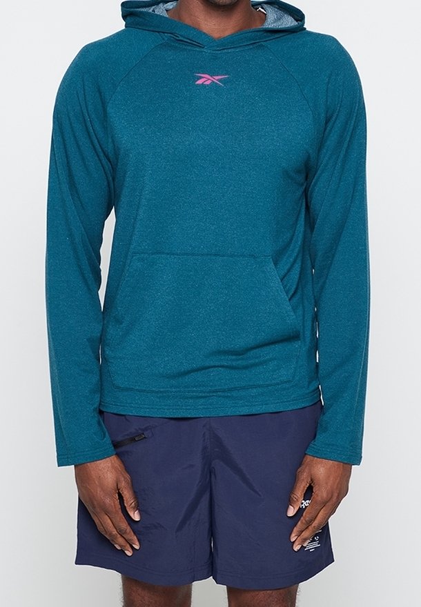 Homme portant un sweat à capuche à manches longues bleu sarcelle avec poche avant et un short de sport bleu marine avec poche zippée, debout devant un fond uni.