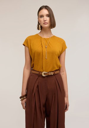 Nainen pukeutunut sinappiväriseen lyhythihaiseen paitaan, korkeavyötäröisiin ruskeisiin laskoshousuihin vyöllä, pitkä kaulakoru, renkaankorvakorut ja rannekoruja yhdellä ranteella.