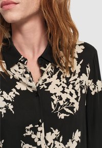 Schwarze Bluse mit einem kontrastierenden blumigen Muster in Beige, mit Kragen und Knöpfen, aus einem glatten Stoff. Nahaufnahme.