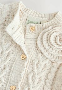 Cremefarvet strikket cardigan med kabelmønstre, en roseformet accent og gyldne hjerteformede knapper. Indeholder et mærkebånd syet indeni.