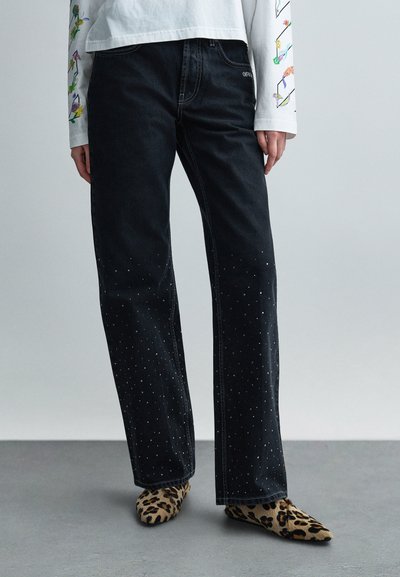 Black denim pants with studded detailing on the lower legs, paired with leopard print slip-on shoes and a long-sleeve graphic top.