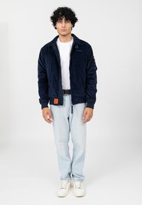 Marineblauwe corduroy jas met een kraag, ritssluiting en zijzakken, gecombineerd met lichtblauwe jeans en witte sneakers. Minimalistisch ontwerp.