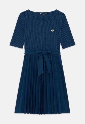 Donkerblauwe jurk met geribbelde top, halflange mouwen, geplooide rok, strik in de taille en klein metalen hartvormig logo op de borst.