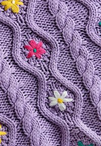 Tejido de lavanda de punto con un patrón de trenzas. Presenta flores bordadas en colores rosa, amarillo y blanco, que añaden color y textura.