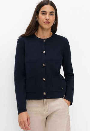 Vrouw met lang donker haar draagt een marineblauwe gebuttonde cardigan met zakken en lichtbeige broek, staande tegen een witte achtergrond.