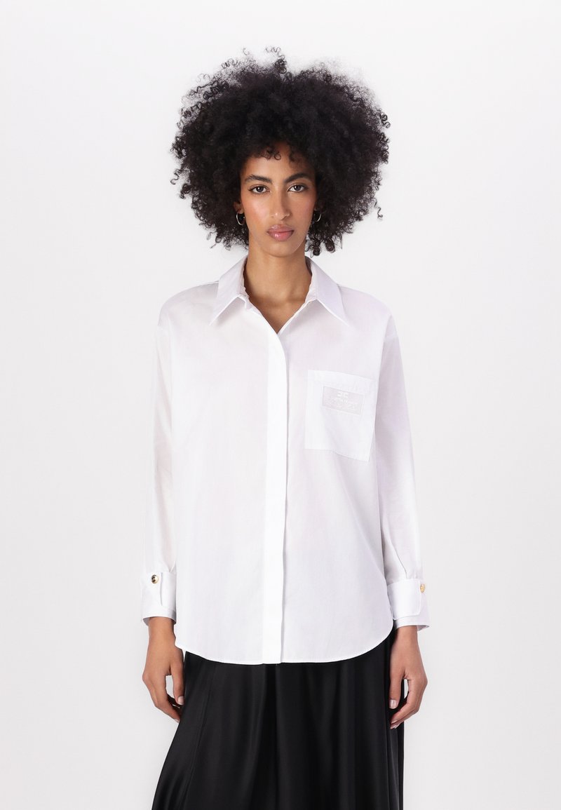 White button-up shirt with a pocket, long sleeves, and gold button accents. Smooth texture and classic collar design.