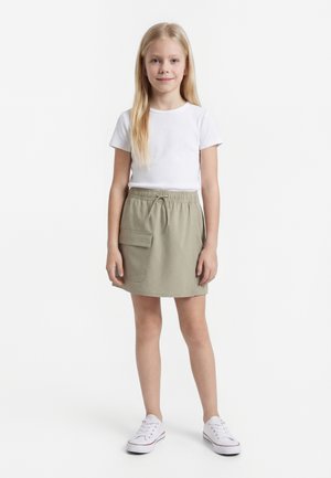 Νεαρό κορίτσι στέκεται, φοράει λευκό κοντομάνικο πουκάμισο, μπεζ φούστα με τσέπη και λευκά αθλητικά παπούτσια σε απλό φόντο.