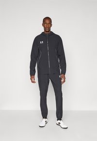 Trening sportiv negru, realizat dintr-un material ușor, cu o jachetă cu fermoar și accente albe, și pantaloni asortați cu manșete elastice.